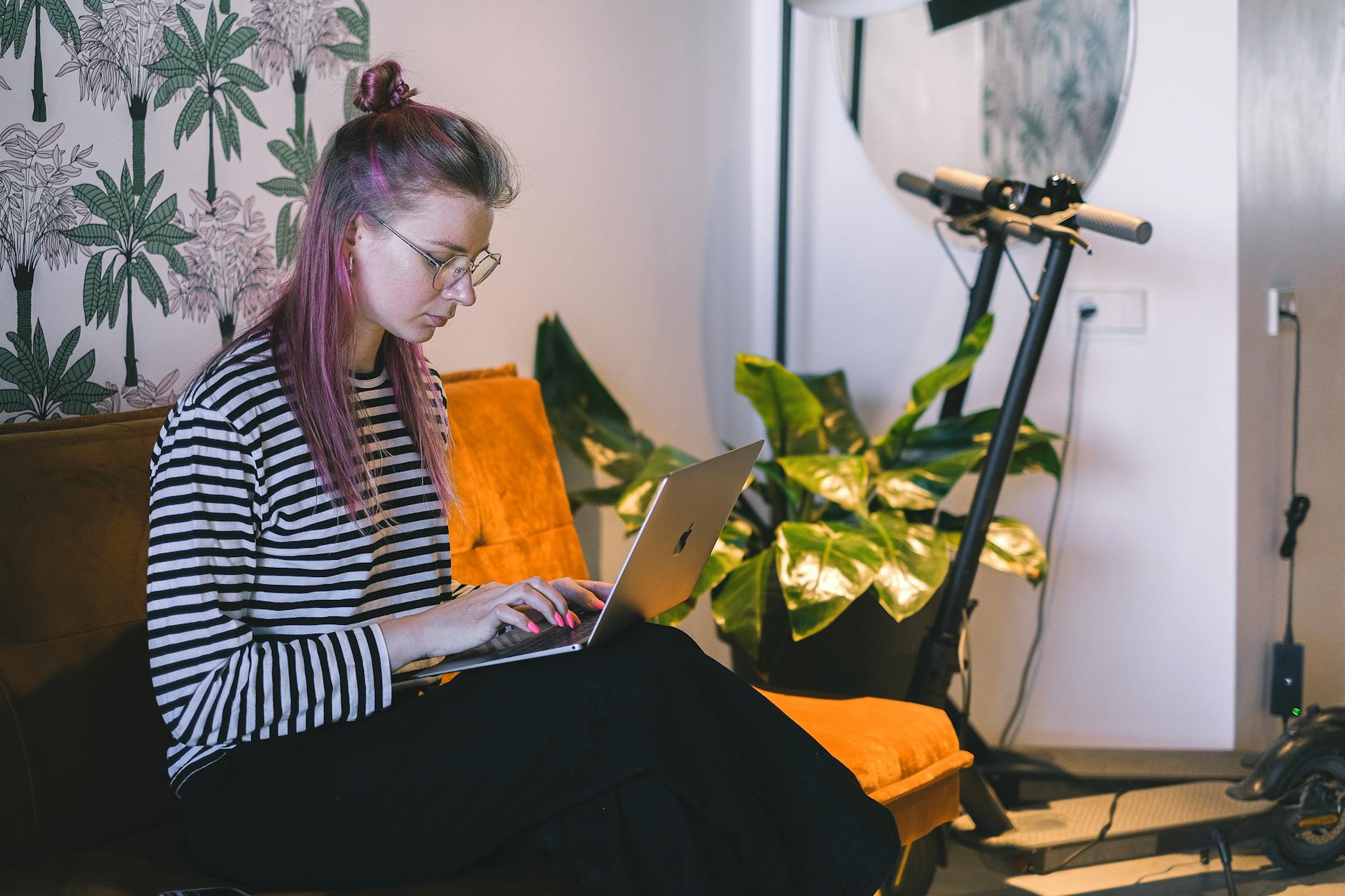 starten met freelancen — A woman with dyed hair types on a laptop while sitting on a couch in a stylish indoor setting with a scooter nearby.
