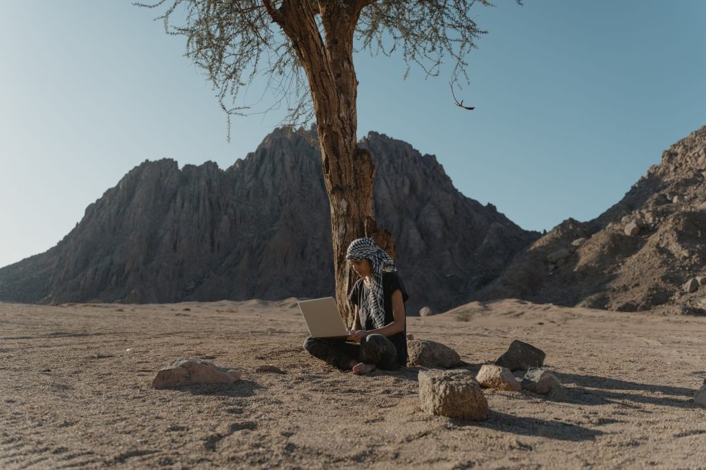 digitale nomaden — Caucasian woman using laptop under tree in rocky desert landscape, embracing remote work outdoors.