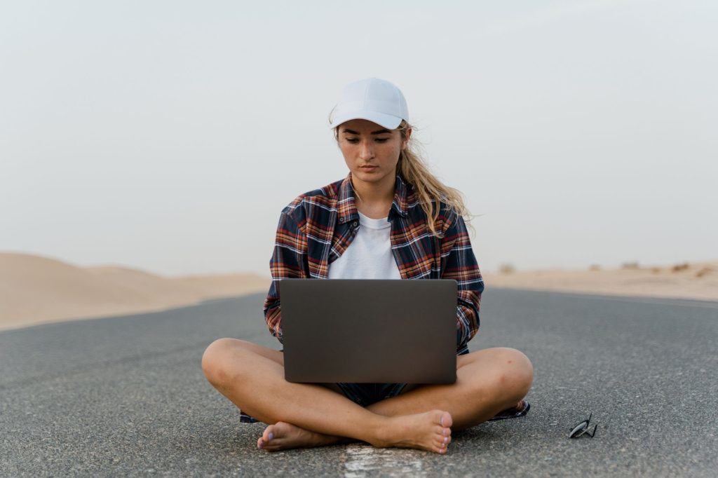 digitale nomaden geld verdienen — Focused young woman sits cross-legged on a road using a laptop, embodying remote work in a desert setting.