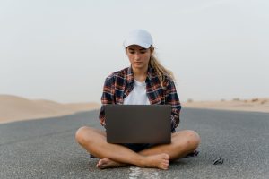 digitale nomaden geld verdienen — Focused young woman sits cross-legged on a road using a laptop, embodying remote work in a desert setting.