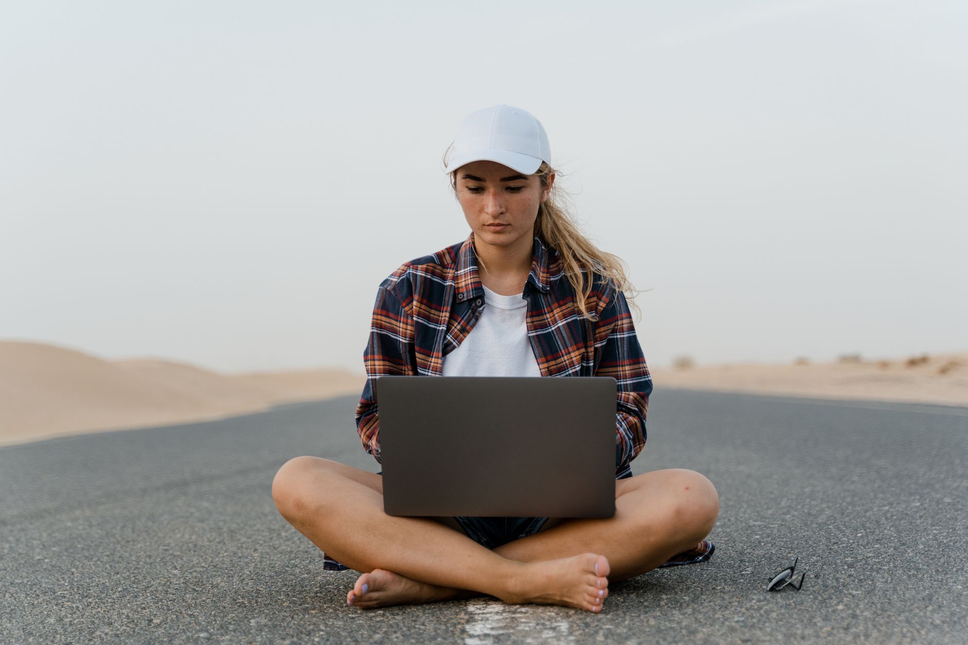 digitale nomaden geld verdienen — Focused young woman sits cross-legged on a road using a laptop, embodying remote work in a desert setting.